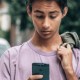 A young person in a purple shirt holds a phone, looking down at it with a jacket draped over one shoulder while standing outdoors, unaware of the potential dangers such as inadvertently accessing pornography on the internet.