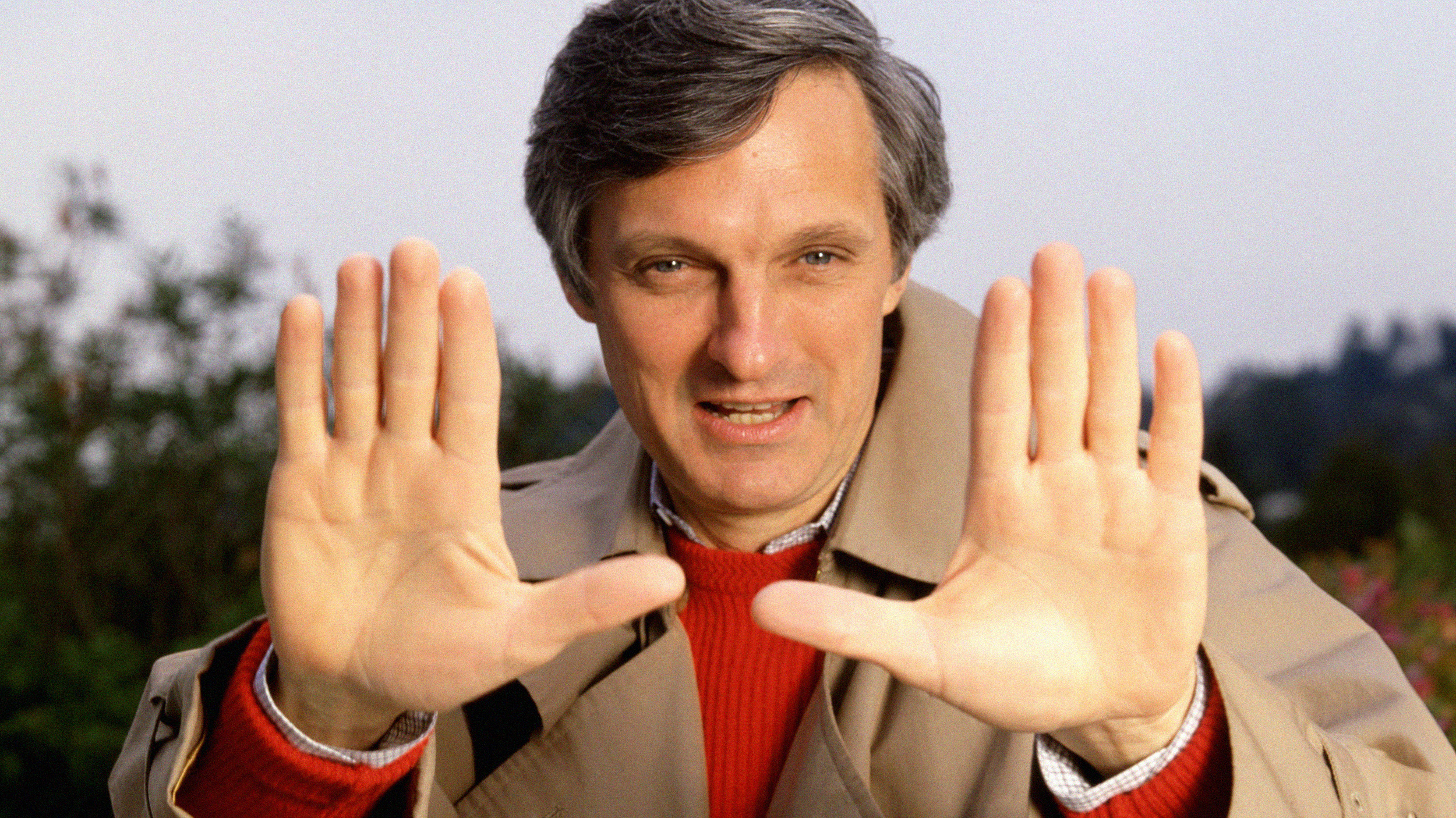 A man in a trench coat, expressing his thoughts clearly with his hands up in the air.