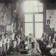 A black-and-white photo of young children sitting in a classroom. Two teachers stand at the back. Drawings and crafts are displayed on the walls, suggesting it was from an earlier historical period, where every good kid learned under watchful eyes.