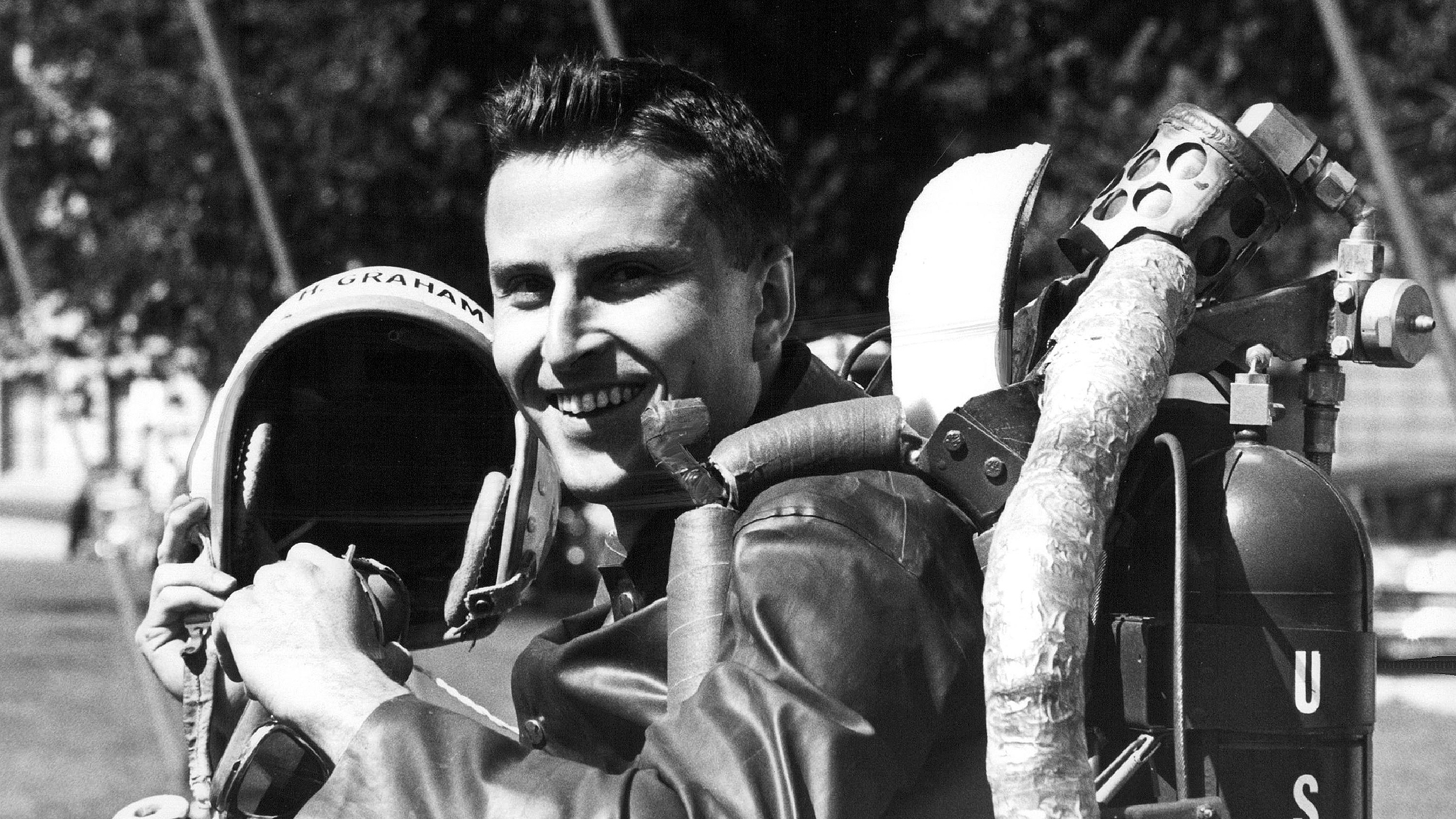 Smiling pilot holding a helmet, wearing a flight suit with a u.s. insignia and a jet pack.