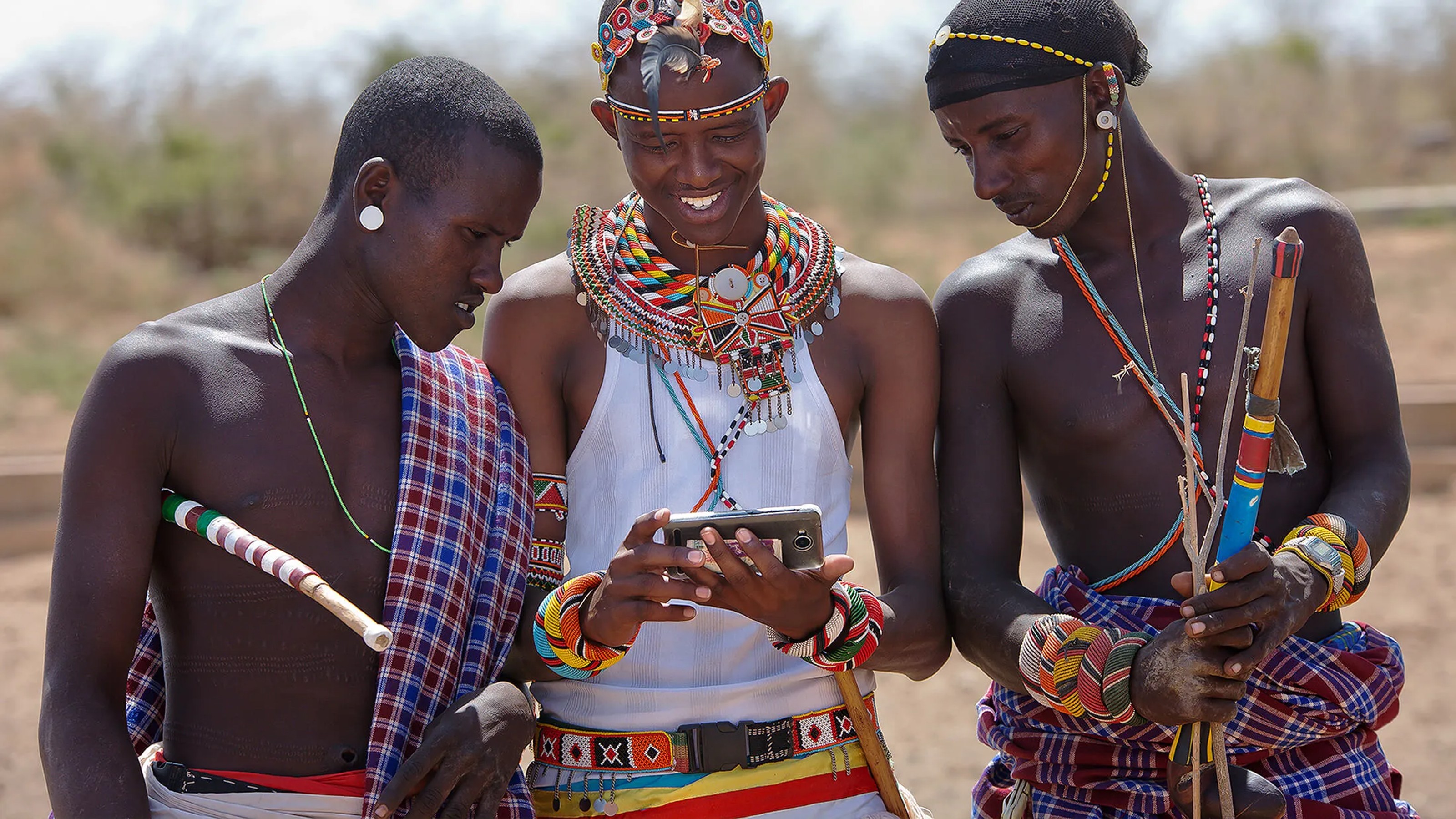 Three Masai men, dressed in traditional attire and adorned with beaded jewelry, gather around a smartphone in an outdoor setting.