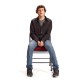 A person with curly hair, reminiscent of Jesse Eisenberg's style, wearing a dark long-sleeve shirt and jeans, sits on a chair against a plain white background.