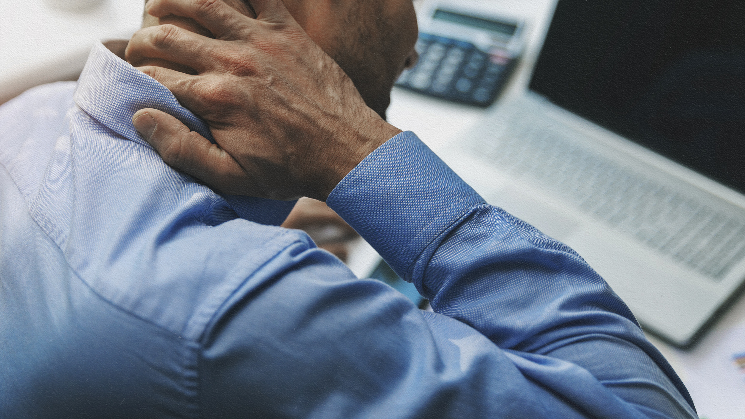 A man in a blue shirt is holding his neck in front of a laptop, possibly experiencing inflammation.