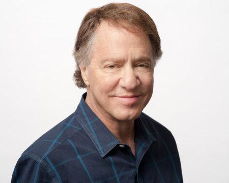 A person with short brown hair wearing a blue checkered shirt, smiling slightly against a plain white background.